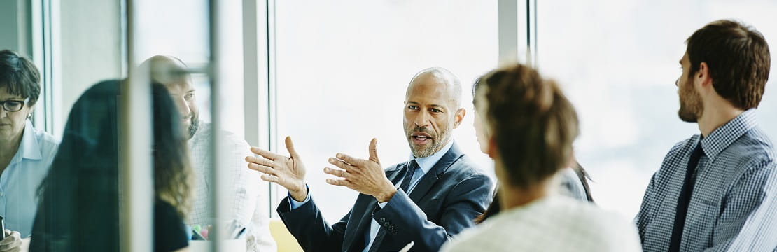 Businessman sits at a table with four other professionals and talks while gesturing with his hands.